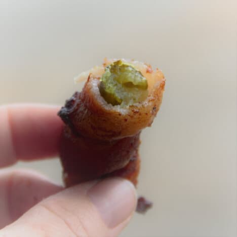 A hand holds up a bacon-wrapped pickle with a bite missing.
