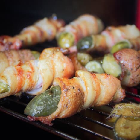 A line of cooked bacon-wrapped pickles sit on a grill.