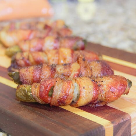 Several cooked bacon-wrapped pickles are arranged on a decorative wooden cutting board.
