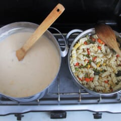 A large saucepan of cheese sauce sitting next to a medium pan of spiral pasta and mixed vegetables on a camp stove.