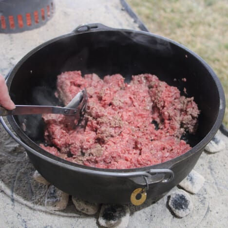 A Dutch oven over orange hot coals with raw ground beef in it.