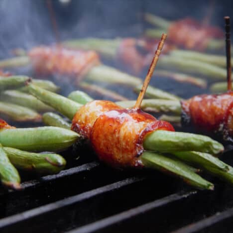 Grilled bacon-wrapped green bean bundles are smoking on a grill.