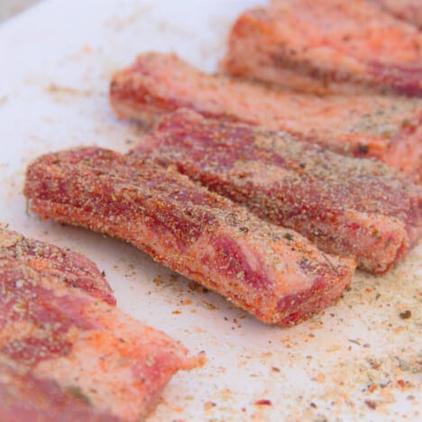 Raw individual lamb ribs sitting on a white chopping board covered in barbecue rub.