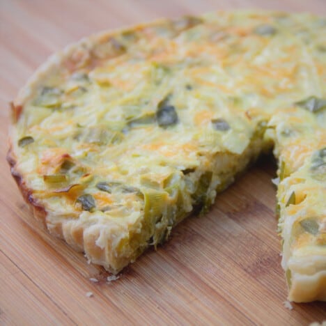 A finished leek tart sits on a wooden cutting board with a slice missing.