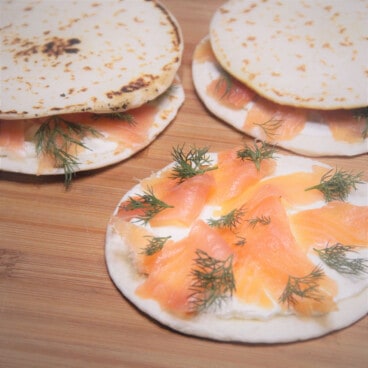 Three quesadillas, one without the top and showing the smoked salmon filling, sit on a wooden cutting board.