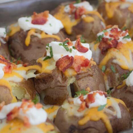 A tray of smashed potatoes topped with melted cheese and garnished with sour cream, bacon pieces and green diced herbs.