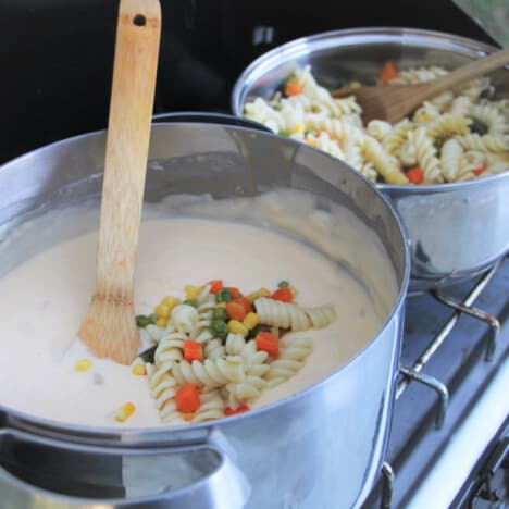 A large pot of white sauce topped with a little pasta and mixed vegetables with the remaining pasta and mixed vegetables in a second pot, both being on a camp gas stove.