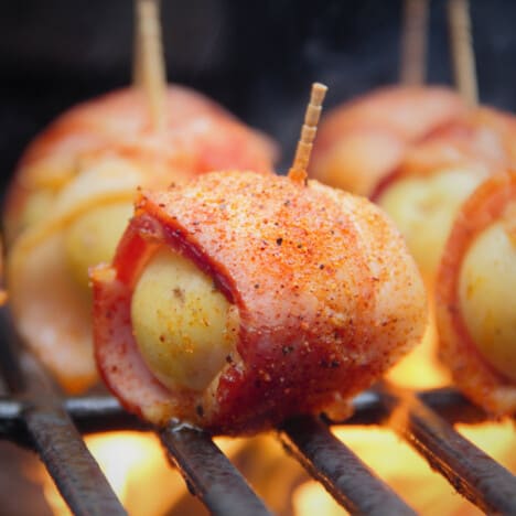 A close up of a bacon-wrapped baby potato on the grill with flames underneath.