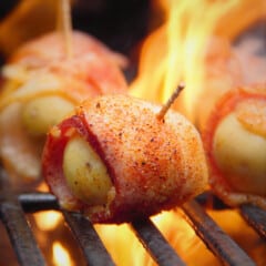 A close up of a bacon-wrapped potato on the grill with flames around it.