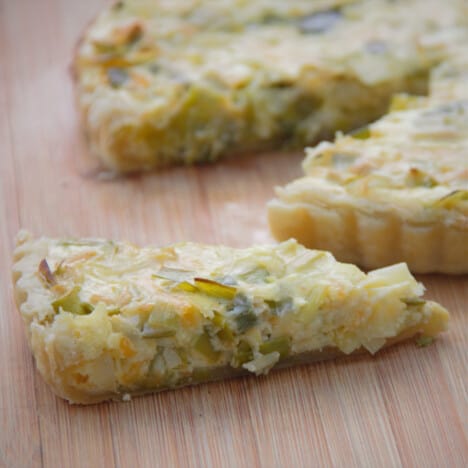 A slice of a bake leek tart sits on a wooden cutting board, with the remaining tart in the background.