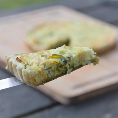 A slice of a baked leek tart held up by a spatula in front of the finished tart in the background.