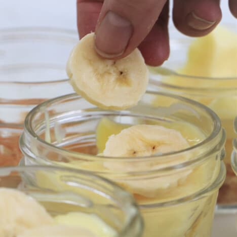 Slices of fresh banana being added to a small jar of banana pudding.