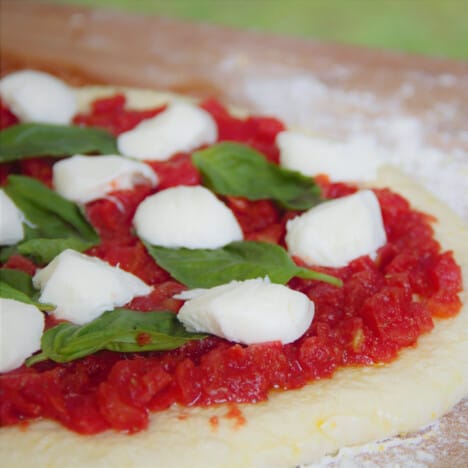 Raw pizza dough on a wooden cutting board is topped with tomatoes, mozzarella cheese, and fresh basil.