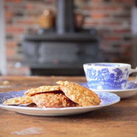 A plate of ANZAC biscuits on an old-fashioned blue plate with a cup and saucer, and wood-fired oven in the background.