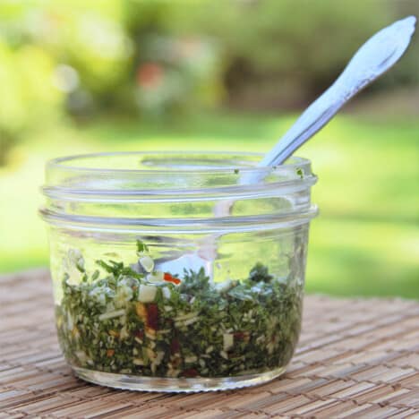 A small glass jar of chimichurri sauce with a spoon in it.