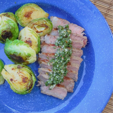 Sliced beef tenderloin topped with chimichurri sauce on a blue camp plate and a side of Brussels sprouts.