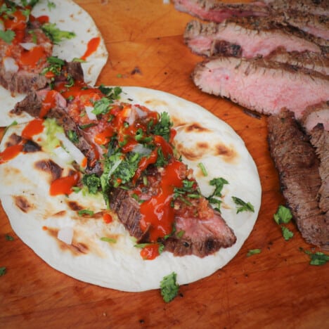 A finished taco sitting on a wooden chopping board sitting next to sliced flank steak.