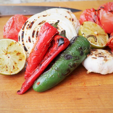 A pile of grilled chilis, tomatoes, onions, and lime on a wooden chopping board.