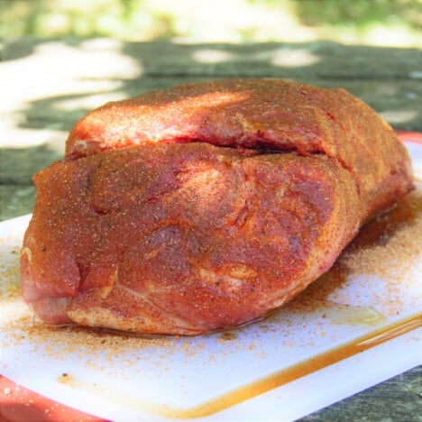 A whole pork butt is covered in rub and resting a white cutting board.