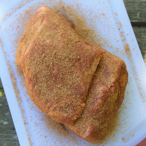 Looking down on a whole pork shoulder butt sitting on a white cutting board, covered in rub.