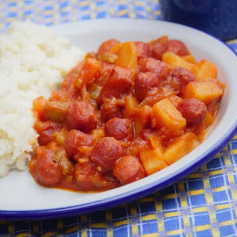 A white camp plate with a serving of sausage stew and side of mashed potato.
