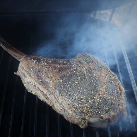 A raw seasoned tomahawk ribeye steak sitting in smoke in a grill.