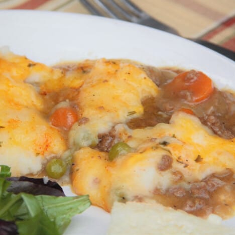 A serving of camp cottage pie sits on a white plate with a side of salad.