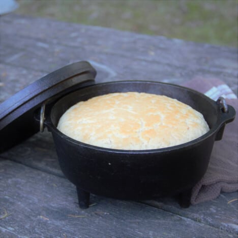 A cooked beer bread sits on a Dutch oven a wooden picnic table.