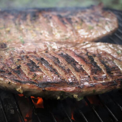 Two flank steak over charcoals with clear grill marks on them.