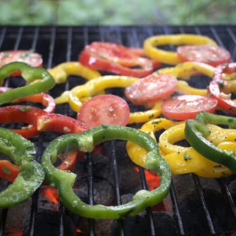 Flat slices of red, gree, and yellow bell peppers along with tomatoes cooking on a grill.