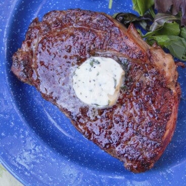 Looking down on a Three Pepper Ribeye Steak topped with a round of compact butter on a blue camp plate.