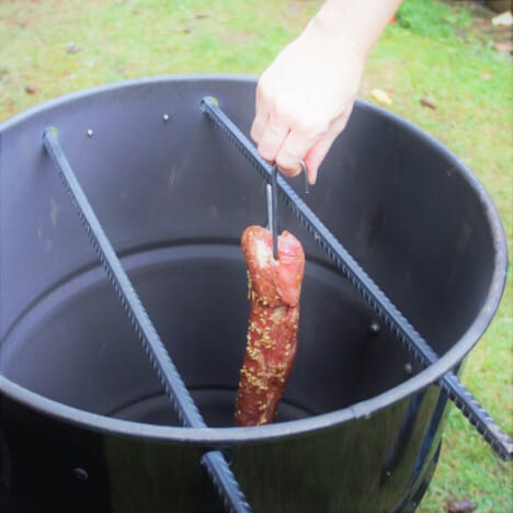 A drum style smoker with a char sui pork tenderloin being hung.