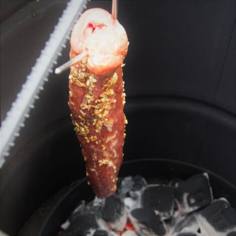 A pork char sui hanging in a round drum smoker with the charcoal below it.