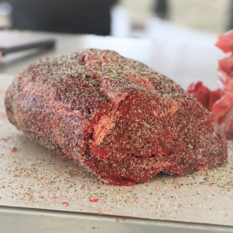 A raw ribeye steak roast sitting on a chopping board covered in pepper and salt.