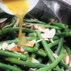 A mustard dressing being poured over green beans topped with sliced almonds.