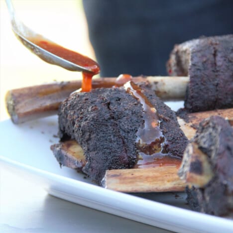 Three beef rib pops on a serving plate with BBQ sauce being drizzled over one of them.
