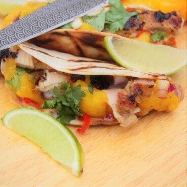 Ready to eat grilled jerk chicken tacos sitting on a chopping board garnished with lemon wedges.