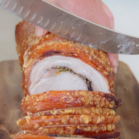 A cooked porchetta with golden skin is being sliced into serving rounds.
