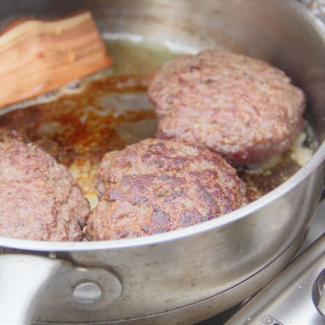 Three kangaroo burger patties cooking in a frypan along with a chunk of wood.