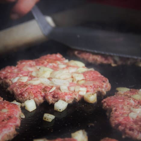 A burger patty, topped with chopped onions, is being smashed on a flat-top grill.
