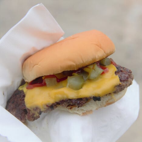 A finished smash burger with cheese and pickles in a bun is wrapped in a white napkin.