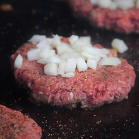 A close up of a unsmashed burger patty with diced onions on a flat-top grill.