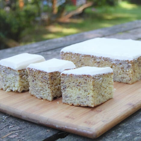 Three squares of banana cake with cream cheese frosting sit on a wooden cutting board.