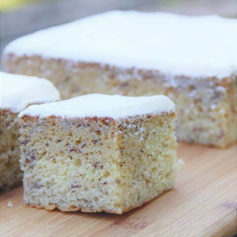 A close up of a square of banana cake with cream cheese frosting on a wooden cutting board.