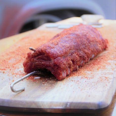 A raw baby back pork rib covered in rub and with the hanging hook added resting on a chopping board.