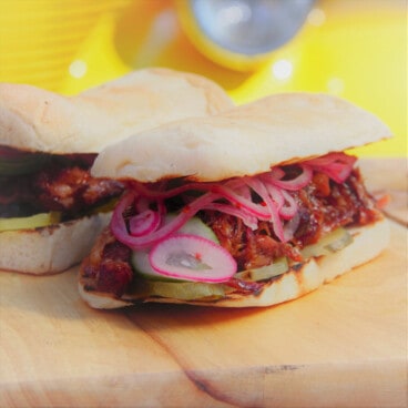A pork rib roll siting on a chopping board with the pickled onions and cucumbers spilling out.