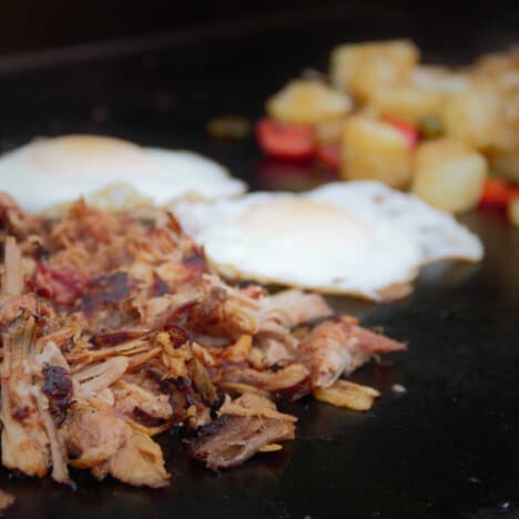 Lines of pulled pork, eggs, and potatoes are cooking on a flat top grill.