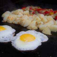 Eggs, potatoes, and bell peppers are cooking on a flat top grill.