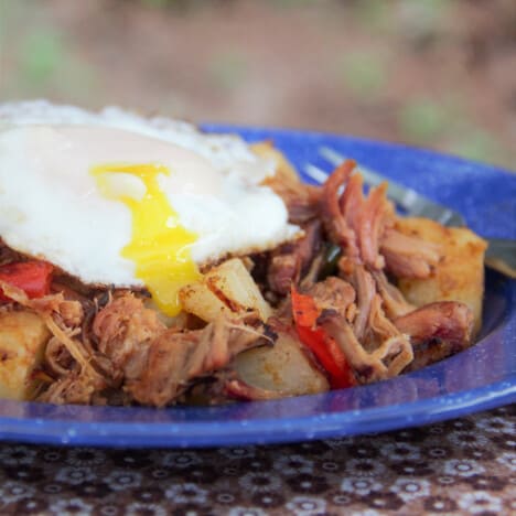 A blue camping plate of pulled pork hash is topped with a runny easy-over egg.