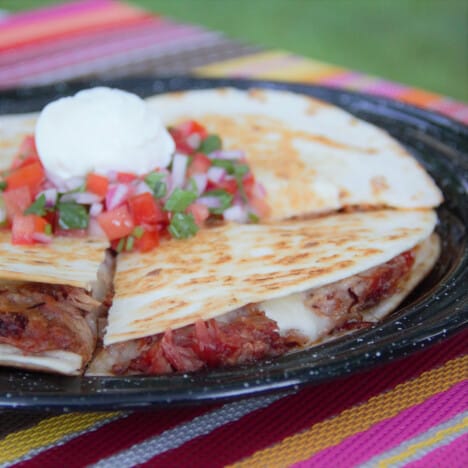 A finished pulled pork quesadilla on a black camping plate, topped with sour cream and pico de gallo.